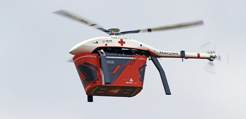White Velos V3 UAV helicopter flying with red cargo payload attached