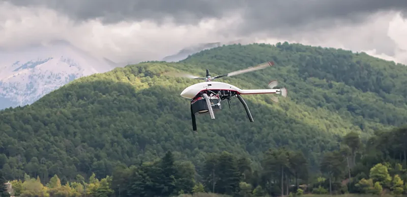 White Velos V3 UAV helicopter flying over green mountain landscape